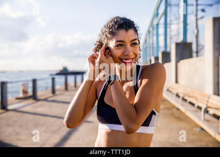 Smiling female athlete adjusting headset while standing on footpath against sky Stock Photo