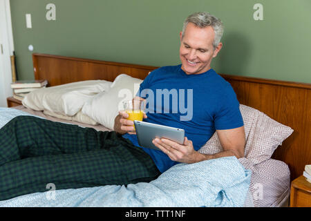 Smiling mature man holding juice while using tablet computer on bed at home Stock Photo