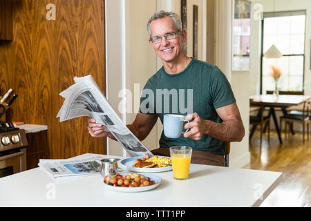 Portrait of smiling mature man holding coffee cup and newspaper while sitting at dining table Stock Photo