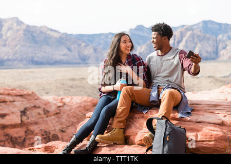 Happy friends talking while taking selfie on rock formations against clear sky during sunny day Stock Photo