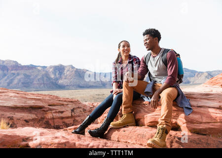 Friends talking while sitting on rock formations against clear sky during sunny day Stock Photo