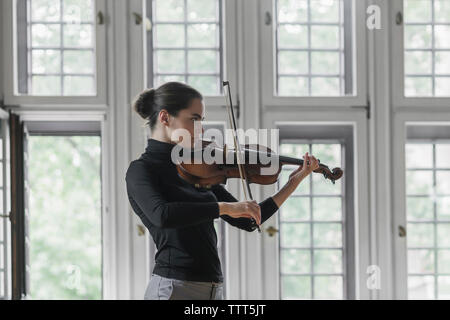 Side view of confident woman playing violin while standing against windows in mansion Stock Photo