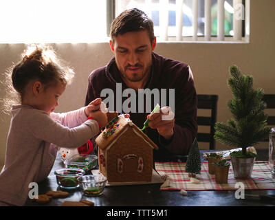Daughter with father decorating gingerbread house on table at home Stock Photo
