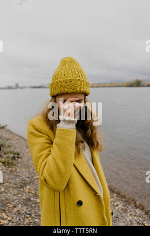 Woman with hand on face wearing winter coat while standing at lakeshore Stock Photo