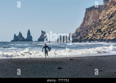 Man with surfboard walking at Black beach against clear sky during sunny day Stock Photo