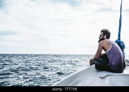 Side view of man looking at sea view while sitting on sailboat against cloudy sky Stock Photo