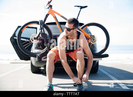 Man with bicycle tying shoelace while sitting on car against clear sky Stock Photo