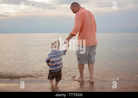 Rear view of father holding son's hand while standing on shore at beach Stock Photo