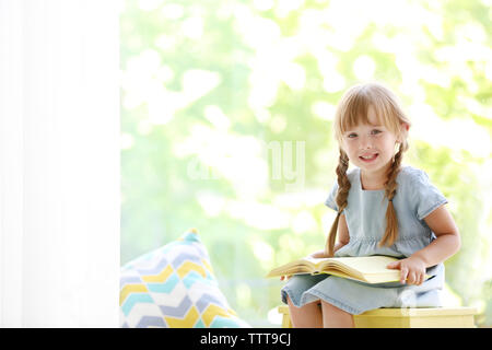 Cute little girl with book sitting on yellow stool Stock Photo