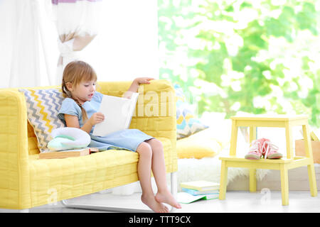 Cute little girl with book sitting on yellow sofa Stock Photo