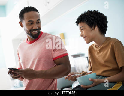 Father using mobile phone while son having breakfast at kitchen counter Stock Photo