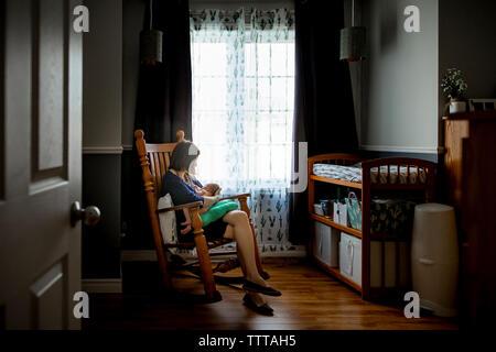 Mother breastfeeding newborn son while sitting on rocking chair seen through doorway at home Stock Photo