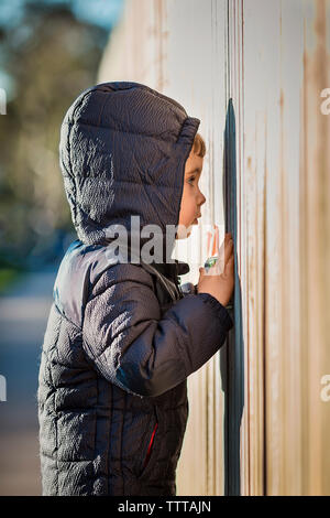 Side view of curious boy wearing hooded jacket while peeking through wooden fence Stock Photo