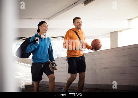 Cheerful male friends holding basketball and skateboard while walking in parking lot Stock Photo