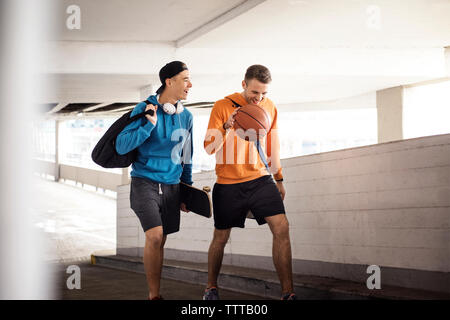 Happy male friends holding basketball and skateboard while walking in parking lot Stock Photo