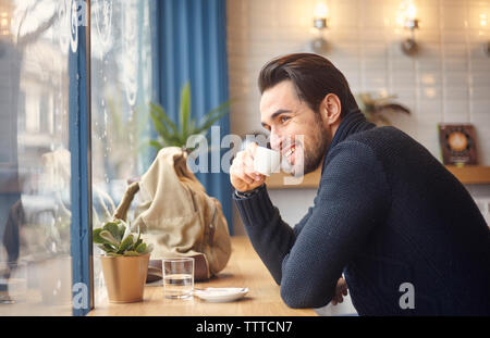 Side view of smiling man looking through window while having coffee by window at cafe Stock Photo
