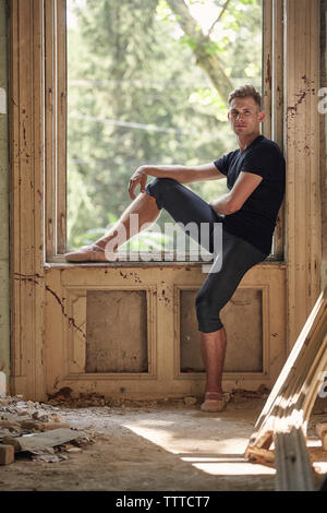 Side view of male ballet dancer sitting on window sill in old building Stock Photo