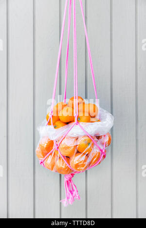 Bunch of fresh oranges in rope basket hanging against wooden wall Stock Photo