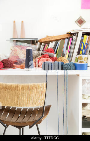 Various threads, yarns and books on table by chair against wall at home Stock Photo