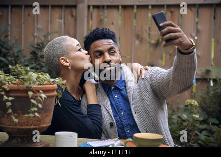 Woman kissing boyfriend while taking selfie in backyard Stock Photo
