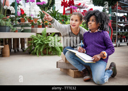 Girl pointing while sitting with friend in plant nursery Stock Photo