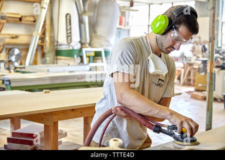 Carpenter using ear and eye protection while sanding wooden plank in workshop Stock Photo