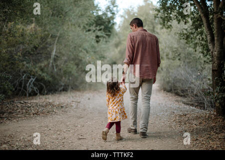 Rear view of father and daughter holding hands while walking on dirt road