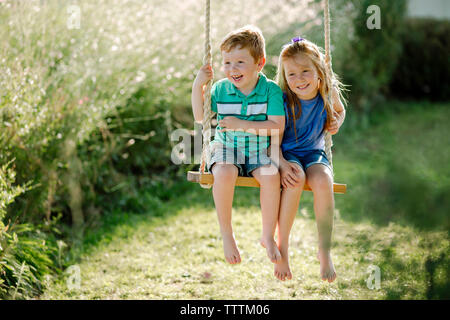 Happy siblings sitting on rope swing at front yard Stock Photo