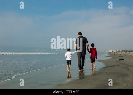 Rear view of father holding sons hands while walking on shore against sky at beach Stock Photo