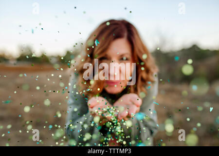 Portrait of smiling woman blowing confetti while standing against sky in forest Stock Photo