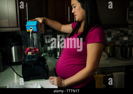 Pregnant woman pouring milk in blender while preparing strawberry smoothie at home Stock Photo