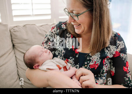 Cheerful mother carrying cute newborn sleeping son while sitting on couch at home Stock Photo