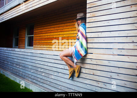 Cheerful woman wrapped in blanket sitting by windows of log cabin Stock Photo