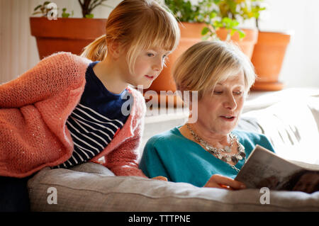 Girl listening to grandmother reading magazine while sitting on sofa at home Stock Photo
