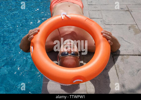 mature bearded man with sunglasses lying next to a swimming pool holding a lifebelt over his head Stock Photo
