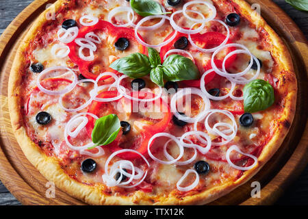 close-up of Freshly baked italian pizza with tomatoes, cheese, onions, ham, olives and bell pepper rings on a wooden round cutting board on a rustic t Stock Photo