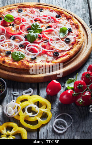 italian pizza with tomatoes, cheese, onions, ham, olives and bell pepper rings on a wooden cutting board on a grey rustic table with ingredients, vert Stock Photo