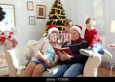 Elderly couple reading book to their grandchildren in living room decorated for Christmas Stock Photo