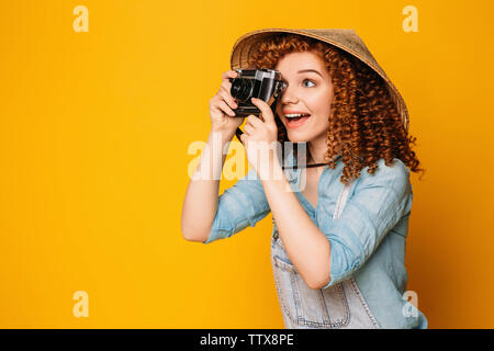 Smiling curly red-haired woman wearing chinese hat holding photo camera and making photo on yellow background. Tourism and travel Stock Photo