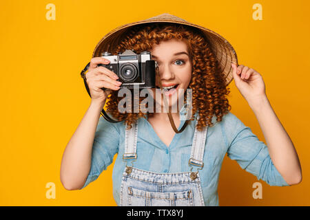 Smiling curly red-haired woman wearing chinese hat holding photo camera and making photo on yellow background. Tourism and travel Stock Photo