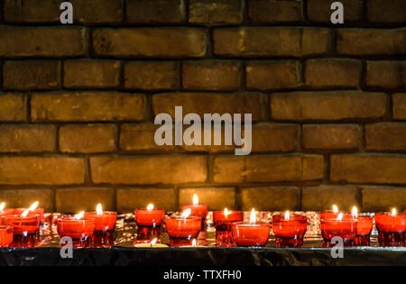 Small white votive candles lighted and placed in red glass holders on a table covered with aluminium foil in front of a brick wall in a church. Stock Photo
