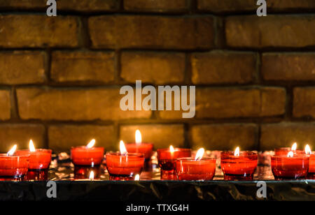 Small white votive candles lighted and placed in red glass holders on a table covered with aluminium foil in front of a brick wall in a church. Stock Photo