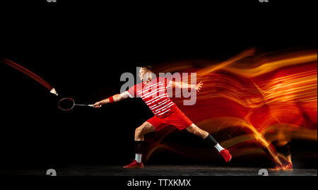 Young man playing badminton isolated on black background in mixed light. Male model with the racket in action, motion in game with the fire shadows. Concept of sport, movement, healthy lifestyle. Stock Photo