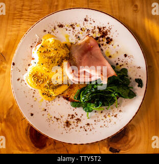 Breakfast meal plate of food comprising eggs Benedict with ham, spinach, toast and garnish set atop wooden table Stock Photo