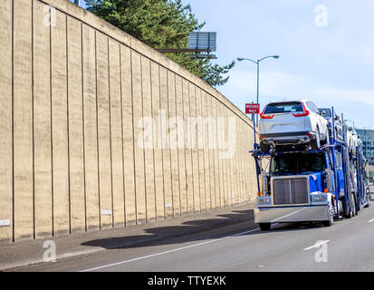 Blue powerful classic big rig car hauler semi truck transporting cars on the special two levels semi trailer running on the road with concrete wall to Stock Photo