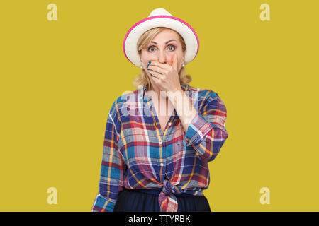 Portrait of shocked stylish mature woman in casual style with hat standing, covering her mouth and looking at camera with unbelievable face expression Stock Photo