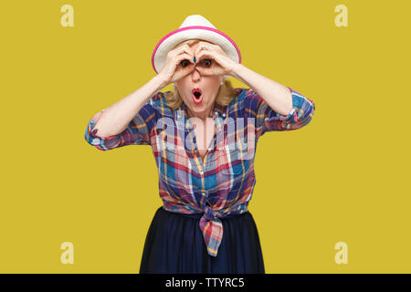 Portrait of funny surprised modern stylish mature woman in casual style with hat standing with binoculars gesture and looking with amazed face. indoor Stock Photo