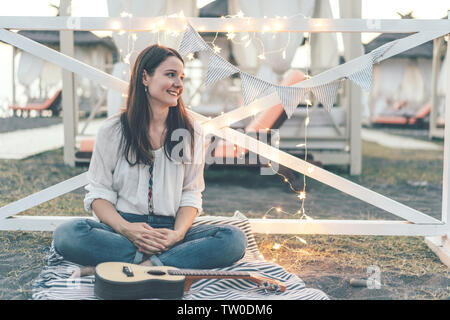 Portrait of a young woman in casual clothing with a ukulele guitar. Evening summer festival with bokeh shiny luminous garlands. Cozy friendly party Stock Photo