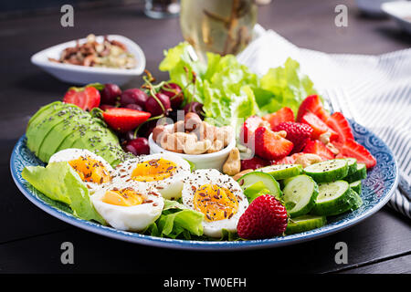 Plate with a paleo diet food. Boiled eggs, avocado, cucumber, nuts, cherry and strawberries. Paleo breakfast. Stock Photo