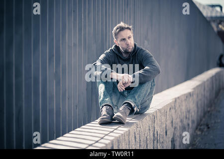 Young attractive man suffering from depression stress sitting alone and sad on the street feeling anxious and lonely in unemployment Mental health Dru Stock Photo